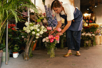 Middle-aged female florist in apron arranging fresh flowers for bouquet in the flower shop. Small...