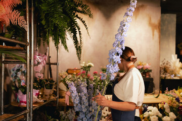 Middle-aged female florist in apron arranging fresh flowers for bouquet in the flower shop. Small...