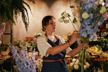 Middle-aged female florist in apron arranging fresh flowers for bouquet in the flower shop. Small...