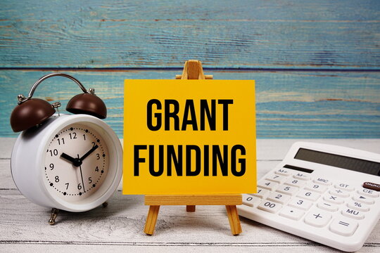 Grant Funding text written on paper card with calculator and alarm clock on wooden background