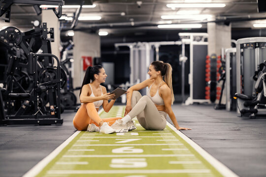 Smiling sportswoman in shape is sitting on gym floor with her personal trainer and discussing progress.