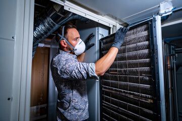 HVAC maintenance. Technician in a patterned shirt, mask, and gloves replacing a large air filter in an HVAC system.