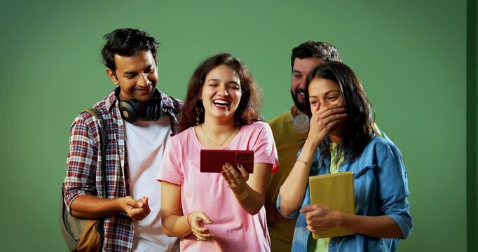 College students watching video on smartphone, Indian university students standing together indoors against green wall enjoying, laughing while looking at funny clip on mobile, joyful moment on camera