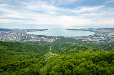 Obraz premium Expansive aerial view reveals Gelendzhik bay with lush greenery in foreground. Cityscape surrounded by hills features prominently under cloudy sky. Serene setting captured in soft daylight