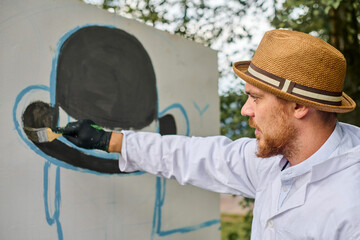 Young male artist in straw hat, white coat painting outdoors. Canvas features abstract figure with hat. Daylight creates natural, calm atmosphere
