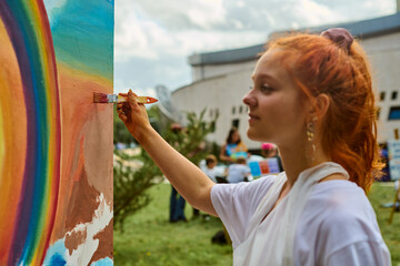 Young female with red hair paints vivid mural outdoors, smiling warmly. Green park, clear sky, vibrant colors create lively atmosphere. Casual attire, relaxed mood
