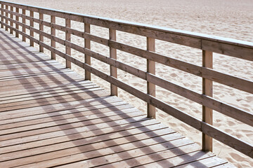 Sunlit wooden boardwalk stretches along serene sandy beach. Shadows from railing create pattern on...