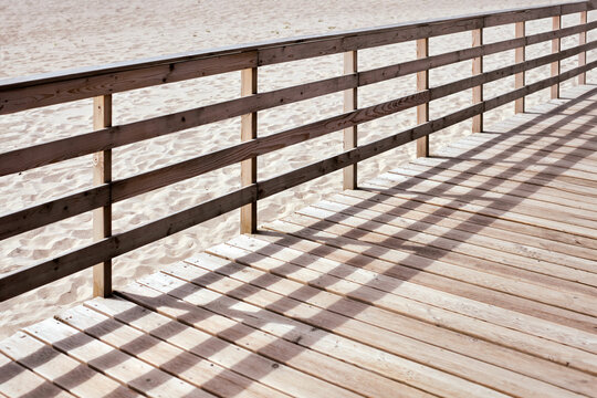 Sunlit wooden boardwalk stretches along serene sandy beach. Shadows from railing create pattern on wooden planks, evoking peaceful and tranquil ambiance
