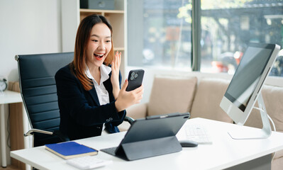 Asian businesswoman in brown blazer working on laptop, analyzing financial data with smartphone. at office