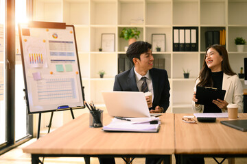 Smiling businessman and businesswoman discuss charts during a relaxed office meeting with coffee, sunlight streaming through the window, and a presentation board beside them