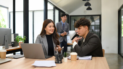 Two young business professionals in suits engage in a focused conversation at a desk with laptops and documents, One-on-One Business Discussion in Modern Office