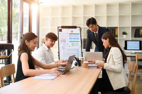 Diverse Business team collaborating during a meeting in a modern sunlit office. A confident leader stands guiding the discussion as colleagues work on laptops and review charts - Powered by Adobe