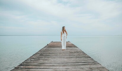 Asian woman standing barefoot on a wooden pier, gazing into the distance over a calm sea horizon, dressed in a white outfit under a pale cloudy sky, mindfulness, wellness retreat, travel blog