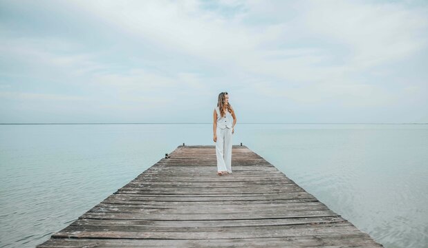 Asian woman standing barefoot on a wooden pier, gazing into the distance over a calm sea horizon, dressed in a white outfit under a pale cloudy sky, mindfulness, wellness retreat, travel blog