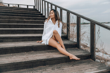 Asian woman sitting barefoot on wooden staircase near lake, gazing into the distance with a relaxed and confident expression, wearing white shirt dress, concept of travel, lifestyle, selfcare