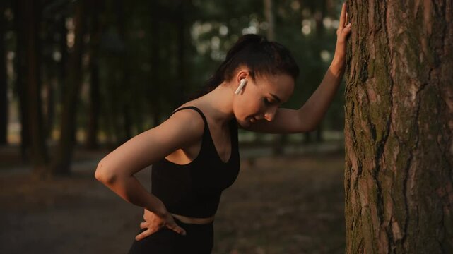 A woman dressed in athletic wear pauses briefly by a tall tree, catching her breath during a rigorous outdoor workout