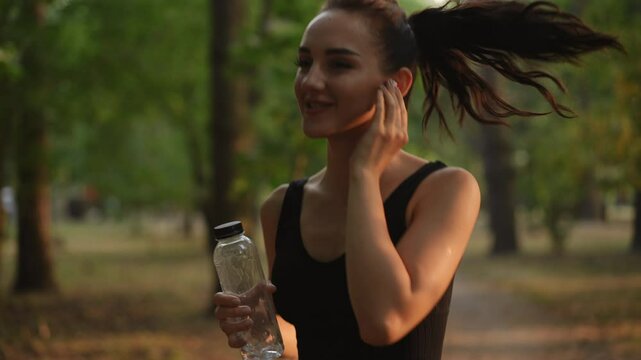 A fit young woman jogs energetically in a park while sipping water and listening to music through earbuds
