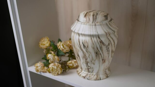 A white vase memorial burn with cremated remains with a brown and white swirl pattern stands on a shelf next to a bouquet of yellow flowers.