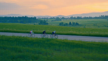 Three helmeted cyclists pedal down a scenic country road, surrounded by lush green fields and...