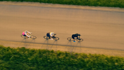 A group of cyclists pedal down a road, their shadows stretching behind them. The aerial view...