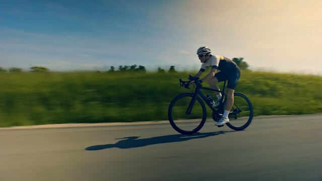 A man, clad in cycling gear and helmet, rides a racing bicycle down a scenic road. The motion blur emphasizes speed and focus.