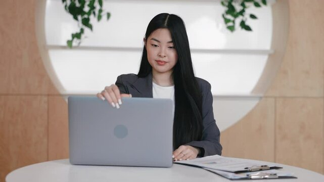 Composed Asian woman lifting laptop lid while preparing for remote work session in tidy office. Getting ready for virtual meeting or starting online tasks while seated beside documents and clipboard.