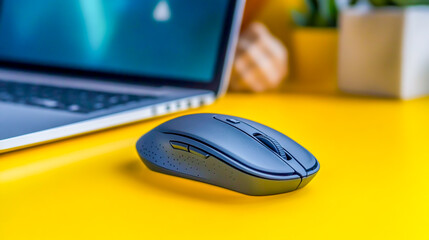 Dark Gray Wireless Computer Mouse on Yellow Surface with Laptop and Decorative Items in Background