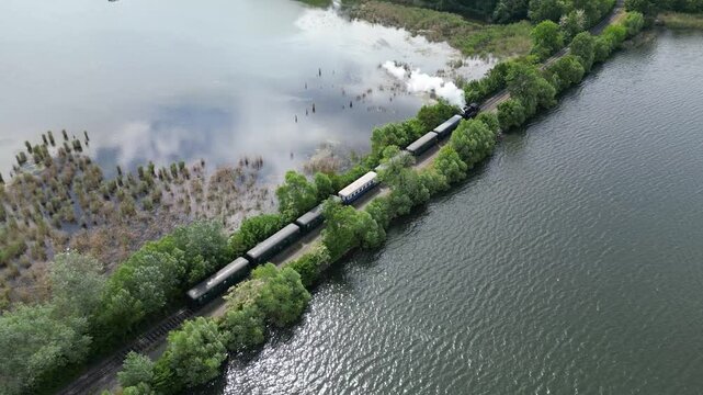 Steam engine train departing lakeside station near Lednice, South Moravia, Czech Republic. Aerial drone spring green trees view. Camera follows the train movement.