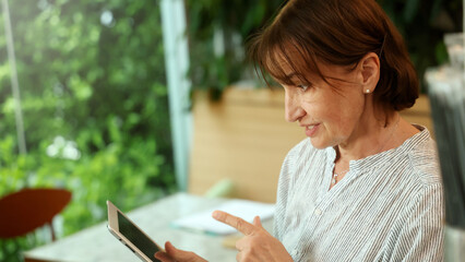 Senior and Technology. Middle aged or senior woman enjoys remote work and online communication technology using various digital devices laptop tablet or smartphone in greeny filled cafe natural light