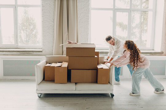 Happy young family couple man and woman push sofa with cardboard boxes in new home. Moving delivery concept.