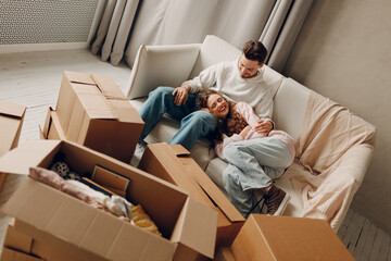 Happy young family couple man and woman relaxing on sofa after moving cardboard boxes to new estate home apartment.