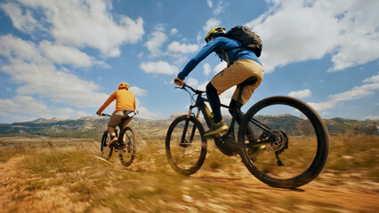 Fototapeta premium Two cyclists pedal through a dirt trail, surrounded by a scenic mountain range. A close-up shot highlights their speed and the motion of their bicycles.