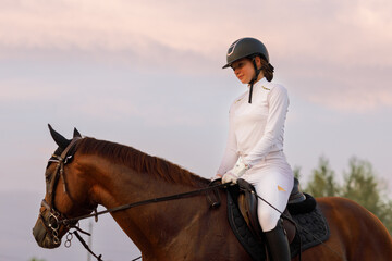 Side view of chestnut horse in harness with female rider jockey in hat helmet and white uniform with bridle stack and boots