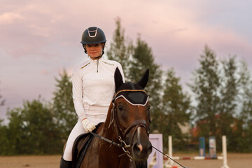 Front view of chestnut horse in harness with female rider jockey in hat helmet and white uniform with bridle stack and boots