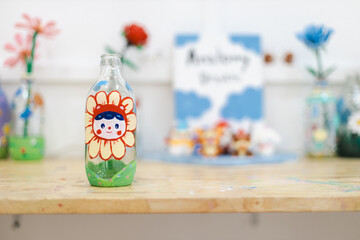 Sustainable ecofriendly craft bottle with painting of smiling flower face art on wooden table surrounded by other creative art pieces in bright room