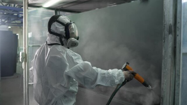 Factory worker wearing protective suit and mask, applying powder coating to metal surface using spray gun