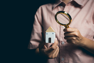 Person Holding Wooden House Model and Magnifying Glass in Dark Background for Conceptual Home...