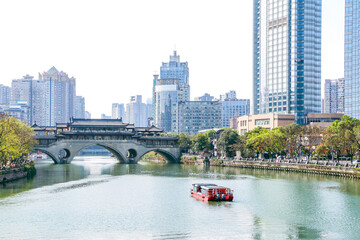 Scenic landscape shot of tour boat on the Jinjiang River about to cross beneath the iconic ancient Anshun Bridge in Chengdu, modern buildings in the backgroung