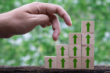 A hand reaches towards stacked blocks with green arrows, symbolizing growth and progress against a natural backdrop.
