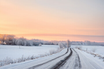 Obraz premium A snowy road curves through a frosty winter landscape at sunrise, with glowing orange skies and trees coated in snow and ice.