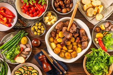 Top view of family dinner table with roasted meat and potatoes in ceramic dish surrounded by fresh vegetables mushrooms cheese olives and bread on wooden surface
