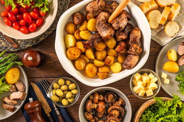 Top view of family dinner table with roasted meat and potatoes in ceramic dish surrounded by fresh vegetables mushrooms cheese olives and bread on wooden surface