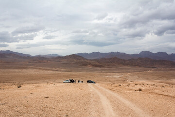 Group of travelers with cars on a remote desert road under a cloudy sky, surrounded by barren...
