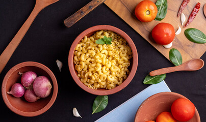 An overhead shot of raw macaroni pasta on the left side of frame,with ingredients placed around it on a  black background. The sharp details enhance its appeal for editorial and marketing use. 
