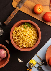 A vertical overhead shot of raw macaroni pasta scattered inside dish,with ingredients placed around it on a  black background. The sharp details enhance its appeal for editorial and marketing use. 