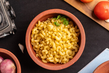 A closeup shot of raw macaroni pasta scattered inside dish,with ingredients placed around it on a  black background. The sharp details enhance its appeal for editorial and marketing use. 