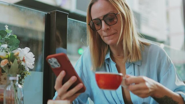 Blonde woman drinking coffee, wearing glasses and blue shirt, working on smartphone at sunny outdoor cafe with floral decor.Travel, tourism, food and drink.