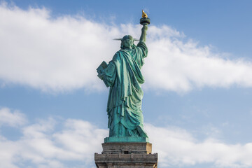 Rear view of the Statue of Liberty with torch raised, seen against a partly cloudy blue sky in New York City. Iconic symbol of freedom and democracy in the United States.