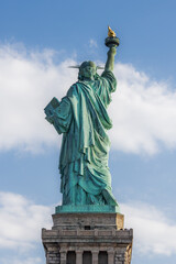 Rear view of the Statue of Liberty with torch raised, seen against a partly cloudy blue sky in New York City. Iconic symbol of freedom and democracy in the United States.