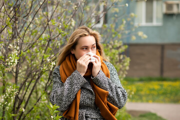 Woman with seasonal allergy gets cold and blows her nose outdoor nearby blooming trees. Healthcare, pollen and flowering allergy in spring concept. World Allergy Day.